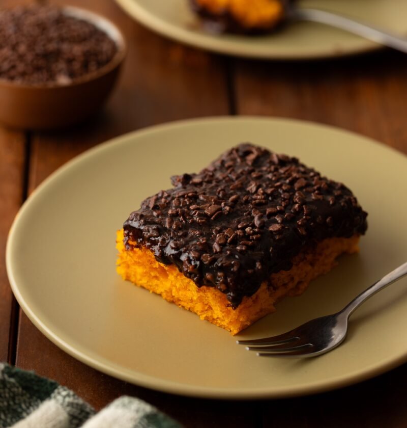 Fatia de bolo de cenoura com cobertura de brigadeiro e granulado ao lado de uma travessa com o bolo inteiro e um pote de granulado de chocolate