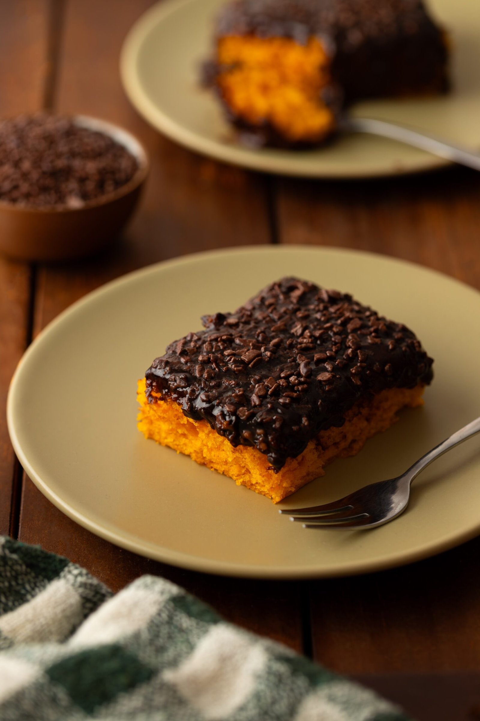 Fatia de bolo de cenoura com cobertura de brigadeiro e granulado ao lado de uma travessa com o bolo inteiro e um pote de granulado de chocolate