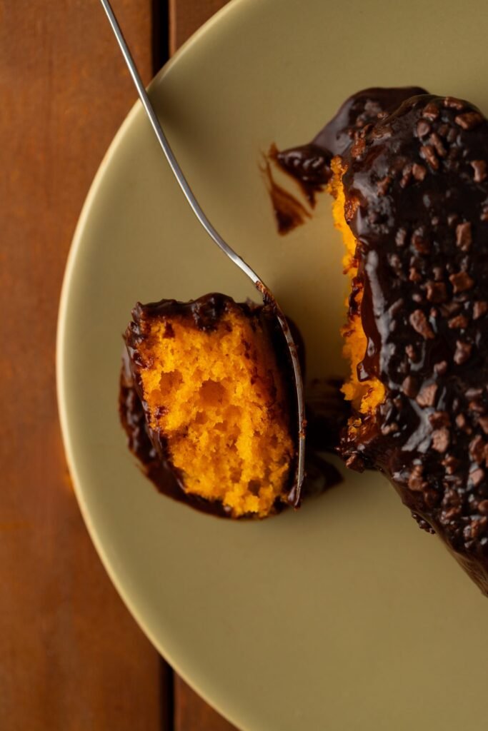 Fatia de bolo de cenoura com cobertura de brigadeiro e granulado ao lado de uma travessa com o bolo inteiro e um pote de granulado de chocolate